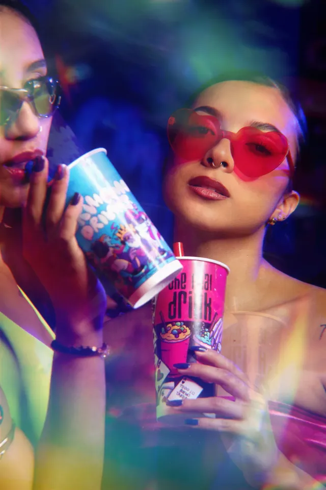 Dos amigas con gafas de sol de corazón sostienen vasos de Hermanos Drinks bajo luces de neón
