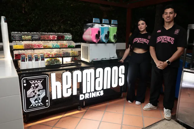 Dos anfitriones de Hermanos Drinks junto al carro móvil con la máquina de granizados y la barra de toppings