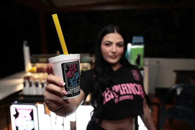 Una anfitriona extiende hacia la cámara un vaso de Hermanos Drinks con pitillo amarillo