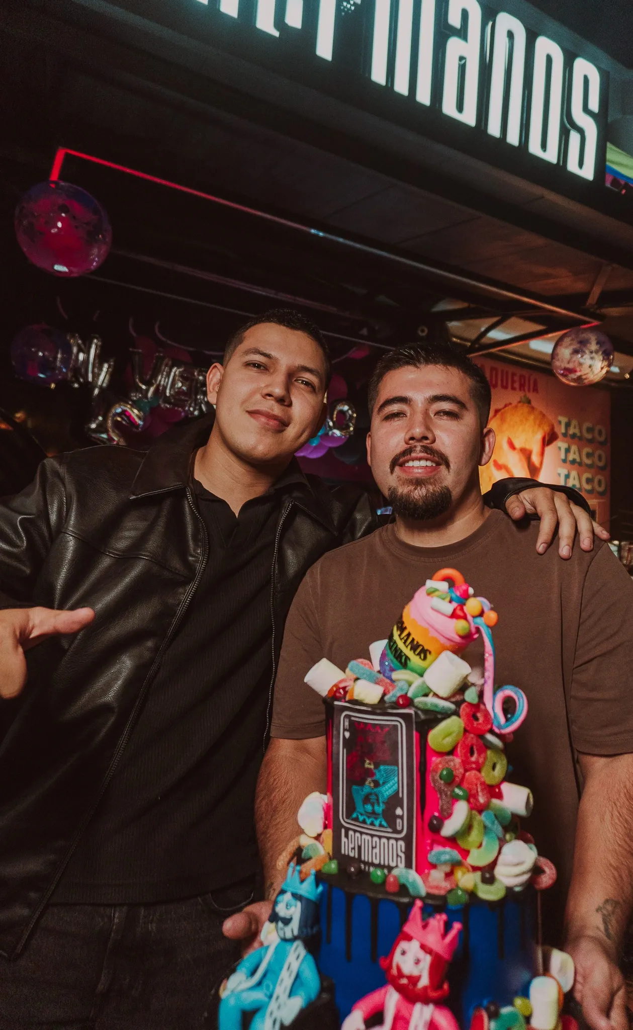 Dos personas posando bajo el letrero de neón de Hermanos Drinks con una torre de dulces coronada por la carta de la marca