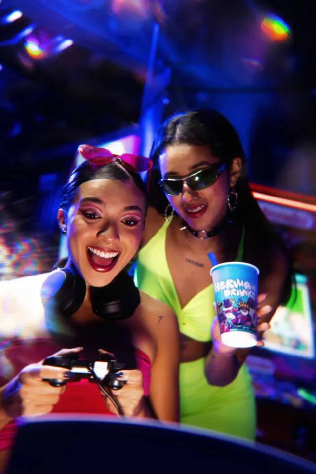 Dos amigas frente a una consola, una con el mando y audífonos al cuello, la otra levantando un vaso de Hermanos Drinks